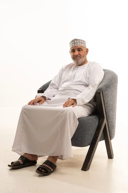 مفهوم الهيبة والوقار، كبير بالسن عماني ،الاهتمام بالمظهر الخارجي والأناقة والتعطر والتطيب, بورتريه لرجل كبير مسن عربي خليجي عماني يرتدي الدشداشة والكمة يجلس على كرسي مريح يقوم بالنظر الى الكاميرا بايماءات السرور و التفائل, خلفية بيضاء