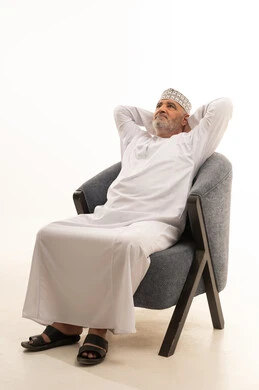 The hands are placed behind the head with gestures of fatigue and contemplation, embodying dignity and respect. An elderly Omani man, concerned with his appearance, elegance, and fragrance, is portrayed. He is an elderly Arab Gulf Omani man wearing a dishdasha and a kumma, sitting on a comfortable chair and looking upwards, with a white background.
