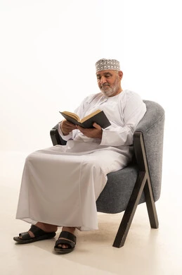 Spending time reading, learning, and gaining culture, acquiring information from books and novels, the concept of self-learning, expressions of contemplation and focus, a portrait of an elderly Arab Omani Gulf man wearing a dishdasha and kumma sitting on a comfortable chair reading a book, white background.