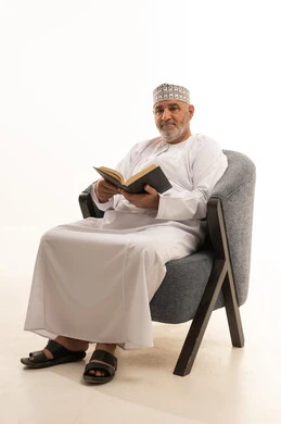 التزود بالمعلومات من الكتب والروايات، قضاء الوقت بالقراءة والتعلم والثقافة، مفهوم التعلم الذاتي، تعابير التمعن والتركيز، بورتريه لرجل كبير بالسن عربي عماني خليجي يرتدي دشداشة وكمة  يجلس على كرسي مريح يقوم بمطالعة كتاب، خلفية بيضاء