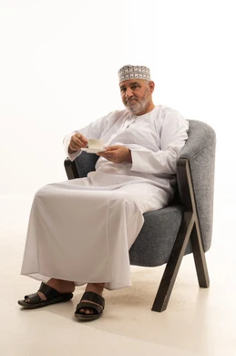 Enjoying hot beverages, looking at something, spending leisure time sipping the favorite drink, a portrait of an Arab Gulf Omani man wearing a dishdasha and kumma sitting on a chair drinking coffee with gestures of happiness and joy, holding on to the noble values inherited from ancestors, white background.