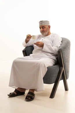 Spending free time sipping a favorite drink, enjoying hot beverages, looking at something, a portrait of an Arab Gulf Omani man wearing a dishdasha and kumma sitting on a chair drinking coffee with gestures of happiness and joy, holding on to the noble values inherited from ancestors, white background.