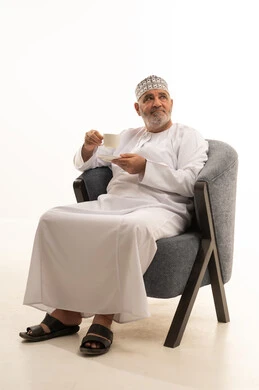 Looking at something, enjoying hot drinks, spending leisure time sipping a favorite beverage, a portrait of an Arab Gulf Omani man wearing a dishdasha and kumma sitting on a chair drinking coffee with gestures of happiness and joy, holding on to the noble values inherited from ancestors, white background.