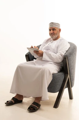 Taking notes and recording information on paper, the concept of self-directed learning, expressions of contemplation and focus, organizing and managing tasks, a portrait of an Omani Arab man wearing a dishdasha and kumma sitting on a chair writing daily tasks and goals, white background.