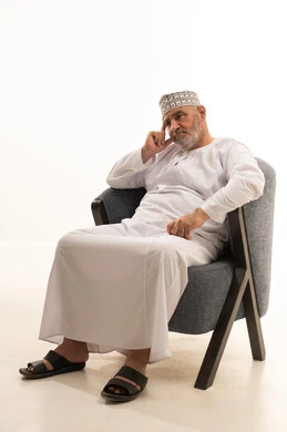 وضع اليد على الراس بايماءات التعب والتفكير، مفهوم الهيبة والوقار، كبير بالسن عماني ،الاهتمام بالمظهر الخارجي والأناقة والتعطر والتطيب, بورتريه لرجل كبير مسن عربي خليجي عماني يرتدي الدشداشة والكمة يجلس على كرسي مريح يقوم بالنظر للأعلى, خلفية بيضاء