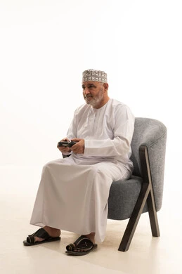 اجهزة وتقنيات الكترونية ترفيهيه حديثة, التفاعل والاندماج مع ألعاب الفيديو، قضاء وقت ممتع ومسلي، بورتريه لشايب عربي خليجي عماني يرتدي الدشداشة والكمة يجلس على كرسي مريح يقوم بالنقرعلى ازرار عصا التحكم، خلفية بيضاء