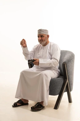 التفاعل والاندماج مع ألعاب الفيديو، اجهزة وتقنيات الكترونية ترفيهيه حديثة,  قضاء وقت ممتع ومسلي، بورتريه لشايب عربي خليجي عماني يرتدي الدشداشة والكمة يجلس على كرسي مريح يقوم بالنقرعلى ازرار عصا التحكم، خلفية بيضاء