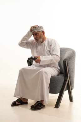 Expressions of sadness for loss, interaction and immersion with video games, modern entertainment electronic devices and technologies, having a fun and entertaining time, a portrait of an elderly Arab Gulf Omani man wearing a dishdasha and a kumma sitting on a comfortable chair clicking on the buttons of a game controller, white background.