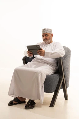 Making use of modern technologies and advanced technology, making video calls through social media applications using a tablet device, a portrait of a smiling Arab Gulf Omani man wearing a kumma and a dishdasha sitting on a comfortable chair, communicating with family and friends, white background.