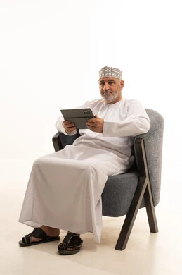 إجراء مكالمات الفيديو عبر تطبيقات التواصل الإجتماعي باستخدام جهاز التابلت،  الاستفادة من التقنيات الحديثة والتكنولوجيا المتطورة، بورتريه لرجل عربي خليجي عماني مبتسم يرتدي الكمة والدشداشة يجلس على كرسي مريح، التواصل مع الأهل والأصدقاء، خلفية بيضاء
