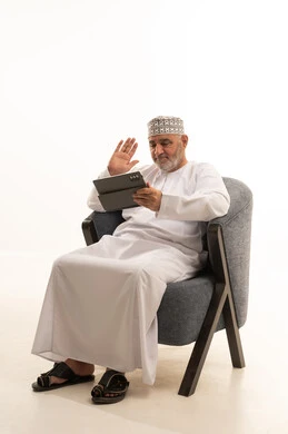 إلقاء التحية والترحيب، التواصل مع الأهل والأصدقاء،  إجراء مكالمات الفيديو عبر تطبيقات التواصل الإجتماعي باستخدام جهاز التابلت،  الاستفادة من التقنيات الحديثة والتكنولوجيا المتطورة، بورتريه لرجل عربي خليجي عماني مبتسم يرتدي الكمة والدشداشة يجلس على كرسي مريح، خلفية بيضاء