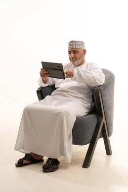 Communicating with family and friends, making video calls through social media applications using a tablet, benefiting from modern technologies and advanced technology, a portrait of a smiling Omani Gulf Arab man wearing a kumma and dishdasha sitting on a comfortable chair, white background.