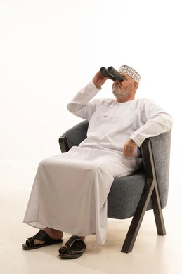 The use of binoculars to view distant objects and bring them closer, a portrait of an elderly Arab Gulf Omani man wearing a kumma and dishdasha sitting on a comfortable chair holding binoculars in his hand and looking at something, a love for exploration and adventure, gestures of enjoyment and pleasure, white background.