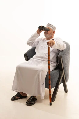 حب الاستكشاف والمغامرة، إيماءات الاستمتاع والسرور، استعمال المنظار للكشف عن الاشياء البعيدة وتقريبها, بورتريه لشايب عربي خليجي عماني يرتدي الكمه والدشداشة يجلس على كرسي مريح يحمل بيده عكاز و المنظار ثنائي العينين وينظر الى شيء ما، خلفية بيضاء .