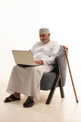 استخدام التقنيات والتكنولوجيا الحديثة للعمل أونلاين، تصفح الانترنت ووسائل التواصل الاجتماعي، انجاز الاعمال والمهام عن بعد عبر الحاسوب المحمول، بورتريه لرجل عربي عماني خليجي كبير بالسن يرتدي الدشداشة والكمه يجلس على كرسي مريح يقوم باستخدام الحاسب، خلفية بيضاء