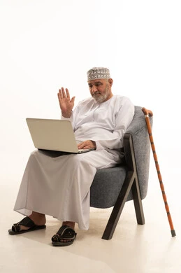 Peace and greetings during communication with family, browsing the internet and social media, completing work and tasks remotely via a laptop, a portrait of an elderly Arab Omani Gulf man wearing a dishdasha and kumma sitting on a comfortable chair using a computer, white background.