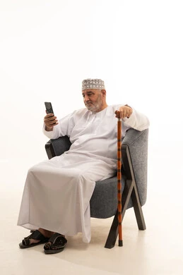 Making a video call via mobile, communicating with family and friends, browsing social media, using modern technology, a portrait of an elderly Arab Gulf Omani man wearing a dishdasha and kumma sitting on a comfortable chair working on a mobile phone with a cane beside him, white background.