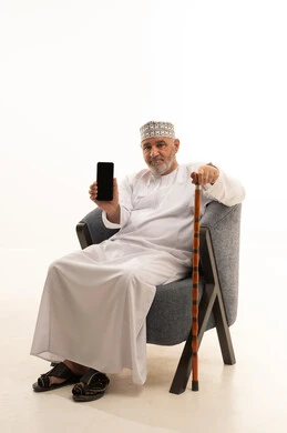 Black mobile screen, using a modern technical device, communicating with family and friends, browsing social media, a portrait of an elderly Arab Gulf Omani man wearing a dishdasha and kumma sitting on a comfortable chair working on a mobile phone with a cane beside him, white background.