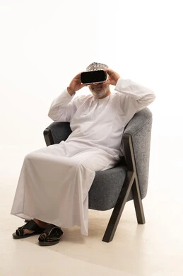 Interaction and integration with holographic elements, the concept of modern electronic technologies, a portrait of an elderly Arab Saudi Gulf man sitting on a comfortable chair wearing a thobe and a headscarf, and 3D virtual reality (VR) glasses, with a white background.