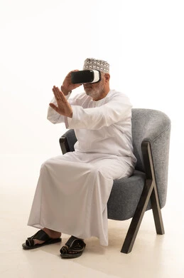 Spending enjoyable and entertaining times with virtual games, the concept of modern electronic technologies, interaction and integration with holographic elements, a portrait of an elderly Arab Saudi Gulf man sitting on a comfortable chair wearing a thobe and a headscarf and 3D virtual reality (VR) glasses, white background.