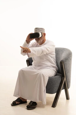 الاستمتاع بمشاهدة أفلام الواقع الافتراضي، مفهوم التقنيات الإلكترونية الحديثة، التفاعل والاندماج مع العناصر الهولوجرامية، بورتريه لرجل كبير بالسن عربي سعودي خليجي يجلس على كرسي مريح يرتدي الدشداشة والكمة ونظارات الواقع الإفتراضي (في آر) ثلاثية الأبعاد، خلفية بيضاء