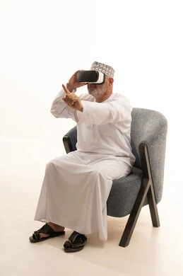 مفهوم الرجولة والوقار،، مفهوم التقنيات الإلكترونية الحديثة، التفاعل والاندماج مع العناصر الهولوجرامية، بورتريه لرجل كبير بالسن عربي سعودي خليجي يجلس على كرسي مريح يرتدي الدشداشة والكمة ونظارات الواقع الإفتراضي (في آر) ثلاثية الأبعاد، خلفية بيضاء