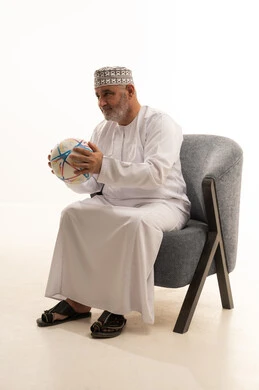 Watching sports matches, excitement and interaction with the national team, attention to appearance and elegance, a portrait of an elderly Arab Gulf Saudi man wearing a thobe and a headscarf sitting on a comfortable chair holding a ball, supporting sports clubs, white background.