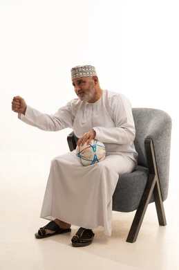 Enthusiasm and interaction with the national team, attention to appearance and elegance, watching sports matches, a portrait of an elderly Arab Gulf Saudi man wearing a thobe and a headscarf sitting on a comfortable chair holding a ball, supporting sports clubs, white background.