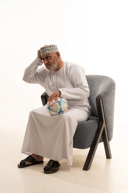 تشجيع الاندية الرياضية، مشاهدة المباريات الرياضية، الحماس والتفاعل مع المنتخب الوطني، الاهتمام بالمظهر الخارجي والأناقة، بورتريه لرجل كبير السن عربي خليجي سعودي يرتدي الدشداشة والكمة يجلس على كرسي مريح  ويحمل الكرة، خلفية بيضاء