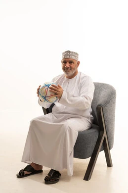 النظر إلى الكاميرا بإيماءات السرور والفوز، الحماس والتفاعل مع المنتخب الوطني، مشاهدة المباريات الرياضية، بورتريه لرجل كبير السن عربي خليجي سعودي يرتدي الدشداشة والكمة يجلس على كرسي مريح  ويحمل الكرة، تشجيع الاندية الرياضية، خلفية بيضاء