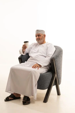 Sitting and enjoying hot drinks and warm winter herbs, looking at the camera with gestures of happiness and joy, spending free time sipping the favorite drink, a portrait of a smiling Arab Gulf Omani elder wearing a kumma and dishdasha sitting on a comfortable chair holding a cup of coffee, blue background.