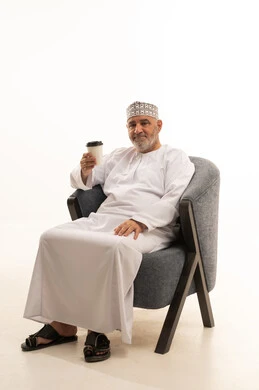 النظر الى الكاميرا بإيماءات السعادة و الفرح, قضاء وقت الفراغ باحتساء المشروب المفضل, الجلوس والاستمتاع بالمشروبات الساخنة والأعشاب الشتوية الدافئة, بورتريه لشايب عربي خليجي عماني مبتسم يرتدي الكمة و الدشداشة يجلس على كرسي مريح يمسك كأس القهوة, خلفية زرقاء
