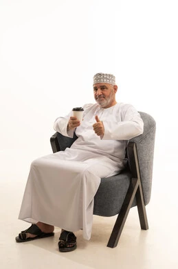 Raising the thumb is a sign of appreciation for the taste of coffee, hot drinks, and warm winter herbs. Looking at the camera with gestures of happiness and joy, a portrait of a smiling Arab Gulf Omani elder wearing a kumma and dishdasha sitting on a comfortable chair holding a cup of coffee, with a white background.