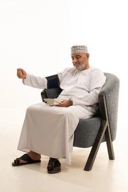 Measuring blood pressure, rapid diagnosis and home care, regular monitoring of general health status, a close-up portrait of an elderly Arab Gulf Omani man wearing a dishdasha and kumma using a medical pressure device to assist himself while sitting on a chair, white background.
