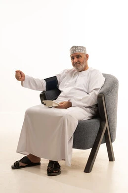 High blood pressure or low blood pressure, quick diagnosis and home care, regular monitoring of general health condition, a close portrait of an elderly Arab Gulf Omani man wearing a dishdasha and kumma using a medical pressure device to help himself sitting on a chair, white background.