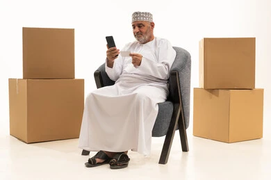 The use of mobile phones in the process of online shopping, a reliable shipping and delivery process, a portrait of an elderly Omani Arab man wearing a dishdasha and kumma sitting on a comfortable chair and working on a mobile phone, the use of modern technological devices, purchasing via the internet, white background.