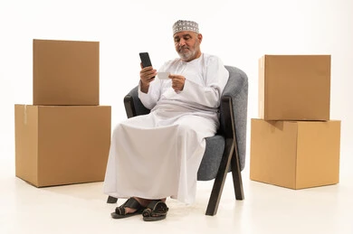 عملية شحن وتوصيل موثوقه, استخدام الجوال المحمول في عملية التسوق الالكتروني ، بورتريه  لرجل كبير مسن عربي عماني يرتدي الدشداشة والكمه يجلس على كرسي مريح ويعمل على الهاتف المحمول، استخدام الاجهزة التقنية الحديثة، الشراء عن طريق الانترنت, خلفيه بيضاء