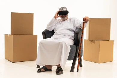Boxes made of brown cardboard, simulating a fantasy world with integration and interaction, a portrait of an Arab Gulf Omani man wearing a dishdasha and kumma and virtual reality glasses sitting on a chair holding a cane, using advanced modern technology and tech, white background.