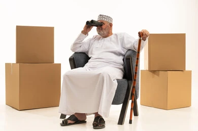 الجلوس على الكرسي بإيماءات التركيز والاستكشاف,  صاديق وكراتين بنية، بورتريه مقرب لرجل كبير بالسن عربي خليجي عماني يرتدي الدشداشة زالكمه يمسك بيده العكاز ويقوم بالنظر عبر منظار التلسكوب ثنائي العينين, رؤية الأشياء البعيدة, خلفية بيضاء