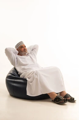 Enjoyment with hands behind the head, the concept of dignity and respect, an elderly Omani man, attention to appearance, elegance, and fragrance, a portrait of an elderly Arab Gulf Omani man wearing a dishdasha and kumma sitting on a comfortable chair looking at the camera with gestures of joy and optimism, white background.