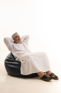 التأمل وشرود الذهن، مفهوم الهيبة والوقار، كبير بالسن عماني ،الاهتمام بالمظهر الخارجي والأناقة والتعطر والتطيب, بورتريه لرجل كبير مسن عربي خليجي عماني يرتدي الدشداشة والكمة يجلس على كرسي مريح يقوم بالنظر الى الكاميرا بايماءات السرور و التفائل, خلفية بيضاء