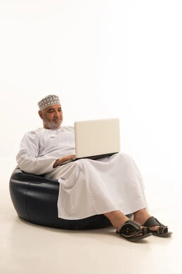 Remote work, using advanced modern technology in professional life, gestures of engagement and focus, a portrait of an Arab Gulf Omani man wearing a dishdasha and kumma working on a laptop while sitting on a comfortable chair, white background.