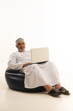 توجيه الوجه نحو الكاميرا بايماءات السرور، العمل عن بعد، استخدام التقنية الحديثه المتطورة في الحياة العملية، ايماءات الاندماج والتركيز، بورتريه لرجل عربي خليجي عماني يرتدي الدشداشة والكمه يقوم بالعمل على الحاسب المحمول يجلس على كرسي مريح، خلفية بيضاء