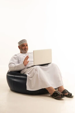 القاء التحيو والسلام عبر مكالمة فيديو، العمل عن بعد، استخدام التقنية الحديثه المتطورة في الحياة العملية، ايماءات الاندماج والتركيز، بورتريه لرجل عربي خليجي عماني يرتدي الدشداشة والكمه يقوم بالعمل على الحاسب المحمول يجلس على كرسي مريح، خلفية بيضاء