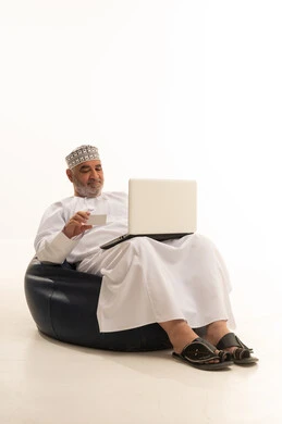 Entering banking information on a laptop, providing means to assist the elderly in the process of online shopping and payment, a close-up portrait of an Arab Omani Gulf man wearing a dishdasha and kumma sitting on a comfortable chair using a credit card for online shopping via the computer, white background.