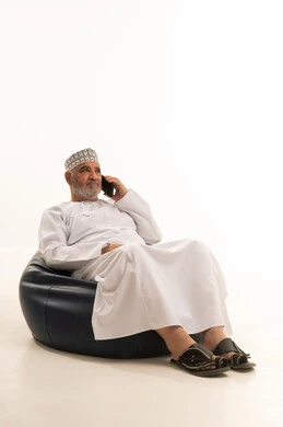 اجراء اتصال هاتفي،  استخدام التقنية الحديثه المتطورة في الحياة العملية، العمل عن بعد عبر الجوال، ايماءات الاندماج والتركيز، بورتريه لشايب عربي خليجي عماني يرتدي الدشداشة والكمه يجلس على الكرسي يقوم بالعمل على الهاتف المحمول، خلفية بيضاء