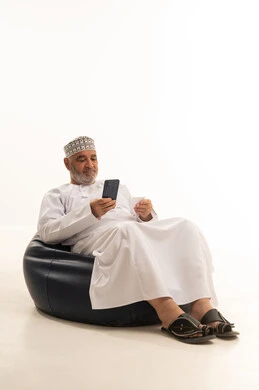 The possibility of paying bills with electronic cards, the ease of purchasing and paying with a bank credit card, a portrait of an elderly Arab Gulf Omani man wearing a dishdasha and kumma sitting on a comfortable chair holding an ATM card and working on a mobile phone, white background.