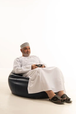مفهوم المتعة والتسلية، إيماءات الاندماج والتفاعل, بورتريه لرجل عربي خليجي عماني يرتدي الدشداشة والكمة يجلس على كرسي مريح يمسك بيده عصا التحكم، قضاء وقت ممتع في اللعب بألعاب الفيديو، الاستمتاع بالتقنيات والتكنولوجيا الحديثة، خلفية بيضاء