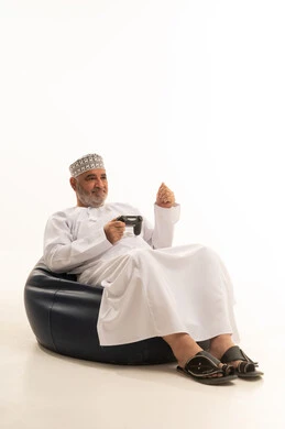 رفع قبضة اليدي بتعابير الفوز، مفهوم المتعة والتسلية، إيماءات الاندماج والتفاعل, بورتريه لرجل عربي خليجي عماني يرتدي الدشداشة والكمة يجلس على كرسي مريح يمسك بيده عصا التحكم، قضاء وقت ممتع في اللعب بألعاب الفيديو، الاستمتاع بالتقنيات والتكنولوجيا الحديثة، خلفية بيضاء