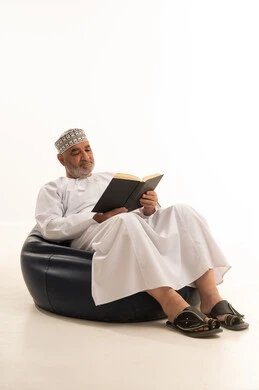 Flipping through and browsing the pages of a book, expressions of contemplation and focus, acquiring information from books and novels, spending time reading, learning, and culture, the concept of self-directed learning, a portrait of an elderly Arab Omani Gulf man wearing a dishdasha and kumma sitting on a comfortable chair reading a book, white background.